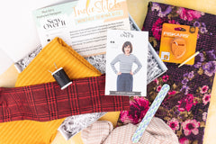 Flat lay of an open IndieStitch box featuring the Roxy Jumper from Sew Over It. Sweater knit fabrics, thread, and an emery board lay on top.