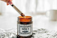 a candle named coffee 'n cobbler sits on a crumpled piece of newspaper. A hand just in frame holds a lighter to the candle's wick.
