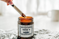 a candle named coffee 'n cobbler sits on a crumpled piece of newspaper. A hand just in frame holds a lighter to the candle's wick.