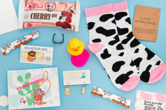 Contents of a western-themed subscription box. Items are cow socks, a cat cowboy pin, a cow girl greeting card, and boot and hat earrings. 