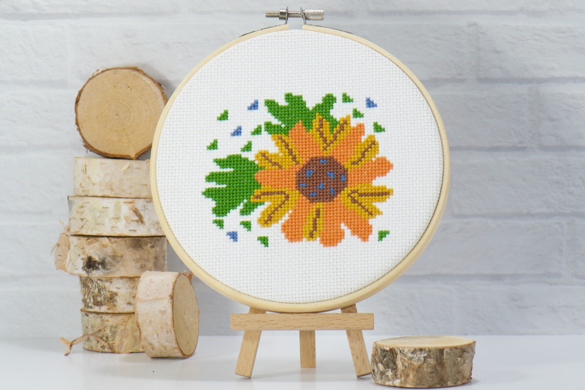 beginner friendly counted cross stitch craft of orange and yellow flower with green leaves on white embroidery fabric inside wood hoop