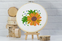 beginner friendly counted cross stitch craft of orange and yellow flower with green leaves on white embroidery fabric inside wood hoop
