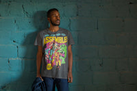Man standing against a blue brick wall and holding a jacket. He is wearing an Assemble Avengers Tshirt.