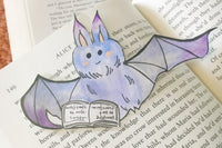 A blue and purple vampire with a sweet smile and blush cheeks bat bookmark reading a book rests on an open hardcover book.