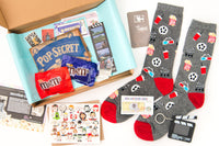 A movie-themed subscription box open surrounded by film reel socks, movie art greeting card, TV pin, clip board keychain, and popcorn.