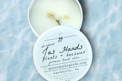 Jas Hands Hand Salve by Greenmade goods. An open tin showing the salve with jasmine buds on top and sitting in a pool of shimmering water.