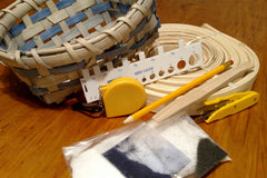 A basketmaking kit showing a completed gray and blue basket, a measuring tape, a pencil, a reed gauge, dye, salt, a reed nipper, more.
