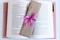 A Romance-themed bookmark is wrapped in brown craft paper and decorated with pink ribbon. It rests on an open hardback book.