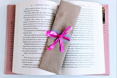 A Romance-themed bookmark is wrapped in brown craft paper and decorated with pink ribbon. It rests on an open hardback book.