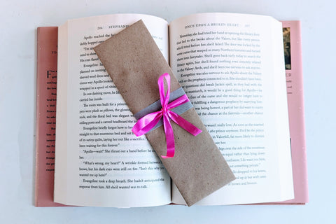 A Romance-themed bookmark is wrapped in brown craft paper and decorated with pink ribbon. It rests on an open hardback book.