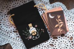 black velvet bag with gold cords embroidered with a bottle and cork next to a deck of cards all on a white lace doily