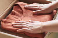 Two hands are resting on a dark orange shirt. Shirt is folded in the box. Left hand has a gold bracelet on. 
