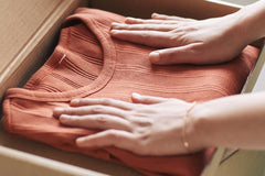 Two hands are resting on a dark orange shirt. Shirt is folded in the box. Left hand has a gold bracelet on. 