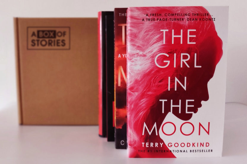 Brown box filled with an assortment of books from various genres, including mystery, romance, and fantasy, featuring vibrant covers.