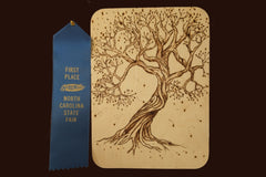 A wood panel with a complex woodburned tree with a first place blue ribbon next to it. The tree seems to be blowing in the wind.