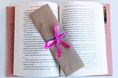A Romance-themed bookmark is wrapped in brown craft paper and decorated with pink ribbon. It rests on an open hardback book.