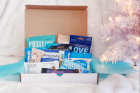 An open, white subscription box filled with healthy snacks. There's a white Christmas tree in the background.