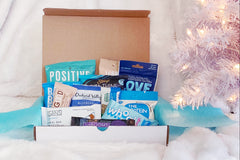 An open, white subscription box filled with healthy snacks. There's a white Christmas tree in the background.