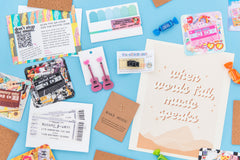 Music-themed box with earrings, art print "when words fail, music speaks" , concert ticket greeting card, and stereo pin on blue background.
