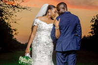 A woman in a wedding dress and man in a tuxedo with arms locked, in front of a landscape scene with the sun setting. 