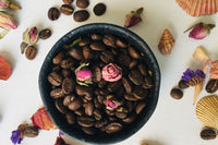 A bowl full of coffee beans with 3 dried roses on top. It is surrounded by seashells, dried flowers and coffee beans.