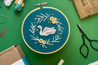 Embroidery project featuring a white swan and blue, white, and yellow flowers on a dark blue fabric, surrounded by embroidery kit supplies.