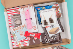 A chocolate-themed subscription box. Items include novelty pen, earrings, holiday greeting card, chocolate spoons, and snacks.