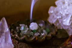 purple and white crystals on either side of photo. a brown bowl with small crystal chunks in the middle with a burning incense cone in the m