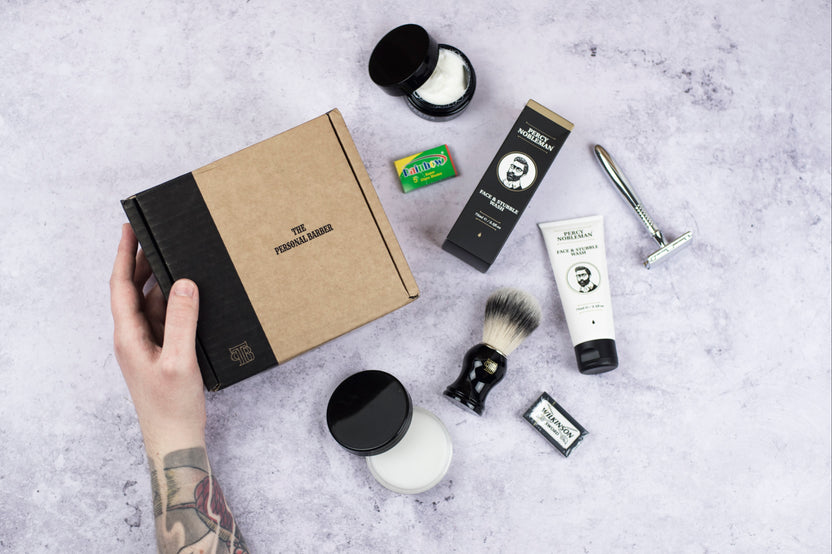A hand hold a closed Shaving Club subscription box with a razor and other shaving products around it.