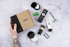 A hand hold a closed Shaving Club subscription box with a razor and other shaving products around it.