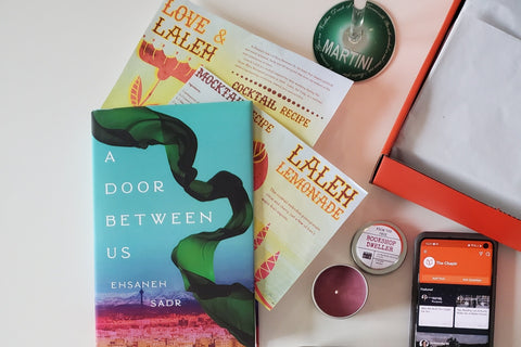 Items from a The Chaptr subscription box including a book, a small candle in a tin, and cocktail recipes.