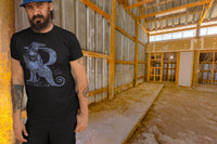 A man leaning on a wall in a barn wearing a Harry Potter Ravenclaw Letter R tshirt. The Letter R is formed by a Raven.