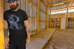 A man leaning on a wall in a barn wearing a Harry Potter Ravenclaw Letter R tshirt. The Letter R is formed by a Raven.