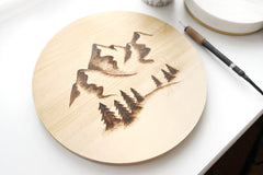 Woodburning project with mountains and forest burned onto a clean cut basswood round sitting next to a woodburning tool.
