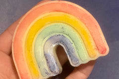 A medium-sized soap shaped and colored like a rainbow (and with textured divots between color spaces) sits across adult-sized fingers,