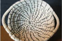 A rope basket made from cotton rope stitched together with waxed thread to make a shallow round basket with a handle on each side.