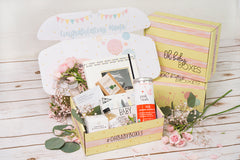 Yellow and pink Oh Baby subscription boxes stacked and one open showing tea, balm, a book and other pregnancy safe products.