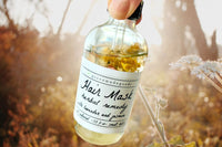 Two fingers holding the Herbal Remedy Hair Mask by Greenmade goods showing dried wild flowers inside the bottle.