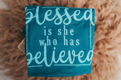 This tee features the uplifting words "Blessed is she who has believed," beautifully displayed against the serene backdrop of teal.