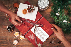 An Artza subscription box with a ribbon around it and lots of Christmas decorations around it. A hand is pulling the ribbon.