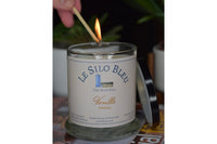 a hand reaching down to light the vanilla candle showing a match and a flame with the candle lid posied on the side of the candle.