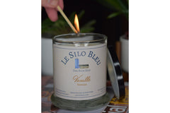 a hand reaching down to light the vanilla candle showing a match and a flame with the candle lid posied on the side of the candle.