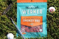 A bag of Werner Smokey BBQ jerky. Lying on the grass surrounded by a golf putter and a Jerky Snob logo golf ball.