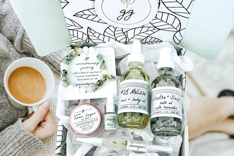 Woman enjoying coffee holding a Greenmade goods Beauty Box. Box includes a bracelet, lip tin, nail pen, mini oil, hair + body oil, bath oil.