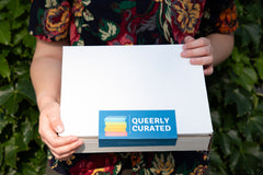 Queerly Curated box (white box, blue label with logo) held by a person with only torso in photo in front of an ivy wall.