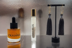 LOKA.HAUS beauty: 'Duality Oil' in amber, center 'Intensity Parfum Roller', & luxe tassel earrings on right. Elegance in a subscription box.