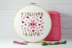 choose love counted cross stitch with hearts and flowers in red, pink, beige, and green on white aida fabric inside wood embroidery hoop.