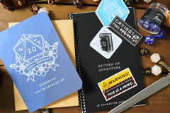 A blue d20 birthday card, a black record of adventure 5e character journal, a glass corked bottle with blue dice, and three other stickers.