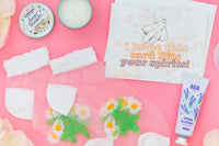 Ghost greeting card with floral background, mini candle open, tube of hand lotion, and sheer daisy flower socks on a pink background. 