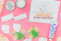 Ghost greeting card with floral background, mini candle open, tube of hand lotion, and sheer daisy flower socks on a pink background. 